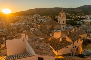 Obraz premium Sunset with golden light in a mountain village with the church tower and the roofs in the center with white facades and the green mountains and the sun with rays in the background and the cloudless 