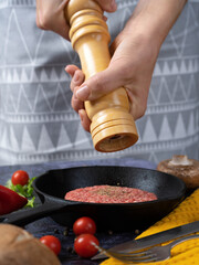 female hands use a wooden mill for spices, pray pepper on a meat patty raw in a cast iron pan