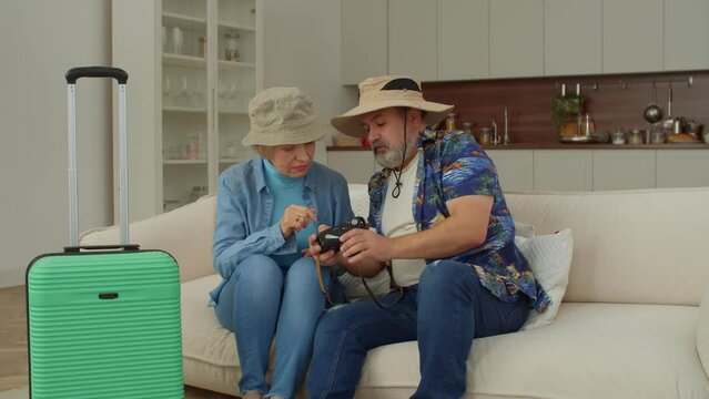 Positive cheerful attractive retired mature traveler couple in sunhats sitting on sofa, watching photos of previous summer trip on digital camera, enjoying good memories and positive emotions.