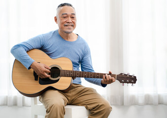 Senior man happy sitting playing acoustic guitar indoors as hobby after retirement. Elderly musician has fun singing a song with an instrument. Cheerful grandfather guitarist enjoys leisure activity.