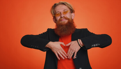 Man in love. Smiling man 20s in black jacket makes heart gesture demonstrates love sign expresses good feelings and sympathy. Young redhead adult guy boy isolated alone on orange studio background