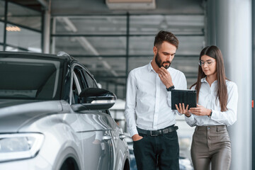Using digital tablet. Man with woman in white clothes are in the car dealership together