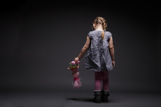 Rear image of a little sad girl with doll, toddler, offended by someone, being in bad mood, over dark grey background.