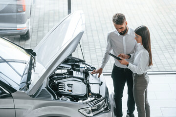 What's under the hood. Man with woman in white clothes are in the car dealership together