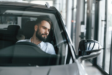 Front view. Sitting inside of automobile. Young man in white clothes is in the car dealership