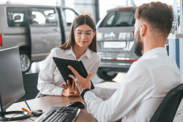 Sitting by table and working. Man with woman in white clothes are in the car dealership together