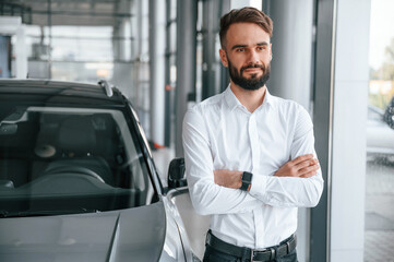Fototapeta na wymiar Standing with arms crossed. Young man in white clothes is in the car dealership