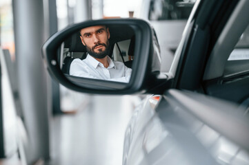 Looking in the side mirror. Young man in white clothes is in the car dealership