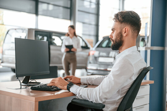 Sitting By A Computer. Man With Woman In White Clothes Are In The Car Dealership Together