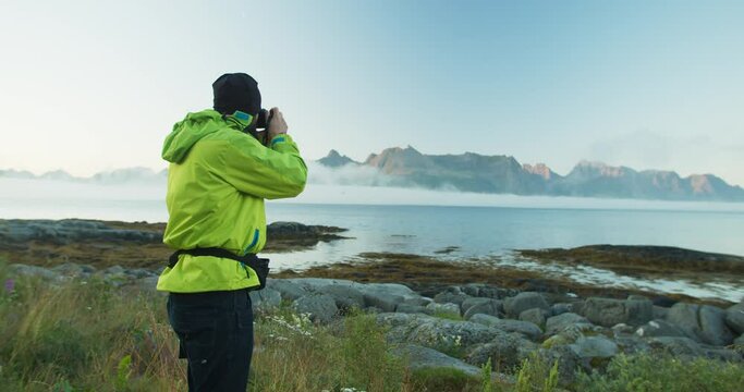 A photographer wearing a bright green jacket takes photos of the beautiful Norwegian fjords. The man is standing with the camera by the lake. Low clouds in the background during the sunny day.
