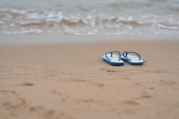 A pair of white  flip flops with blue straps rests on sandy beach with a small wave in front of the sea. Sponge shoes were removed and left in wet sand, and there were blurry images of human footprint