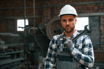 Standing in grey uniform and in white hard hat. Factory male worker is indoors