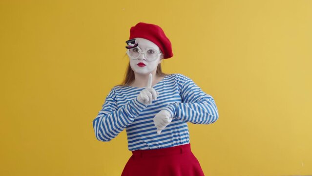 Portrait of female expression mime artist against yellow background. Woman playing a pantomime performance, showing a time on her wrist