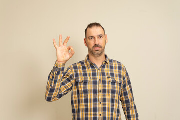 Fototapeta premium Handsome funny smiling man in checkered shirt posing isolated on beige wall background, studio portrait. People candid emotions lifestyle concept. Mock up copy space. Showing OK gesture.
