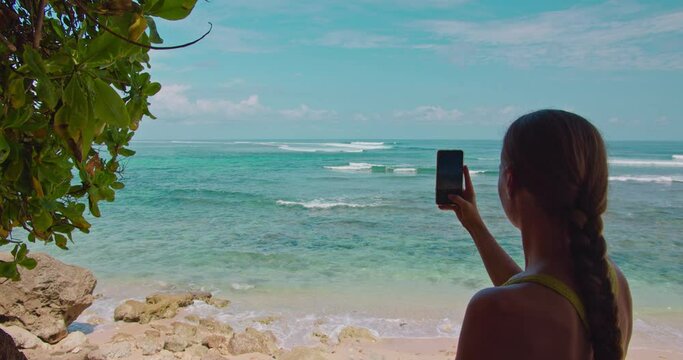 Rear slow motion shot of young woman taking picture of ocean landscape with her smartphone. Tourism, technology, vacation and leisure concept. Bali Island, Indonesia.