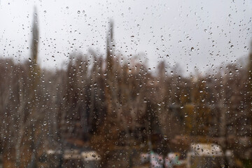 Textured surface of drops of autumn cold rain on window glass against blurred background of an empty dark city park and gray sky. Rainy weather.