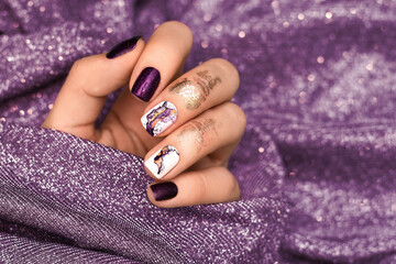 Female hand with purple nail design. Glitter purple nail polish manicure with abstract nail art. Female model hand on glitter purple fabric background
