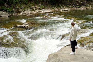 Horizontal image of young stylishly dressed woman near river and rapids looking at nature. Place for your design