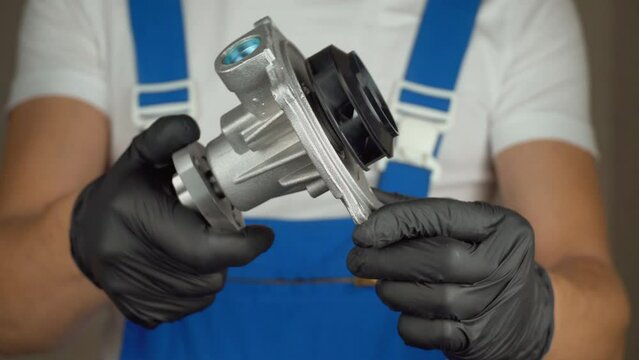 Car mechanic in blue jumpsuit holds water pump for cooling in his hands and rotates it in black gloves. . Concept of replacement of pump in engine in car, automotive spare parts. shows class sign.
