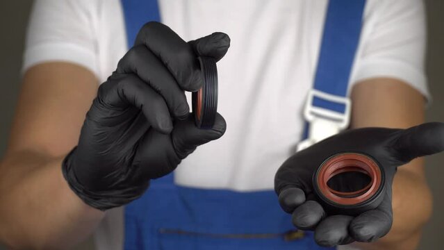 Auto mechanic in blue jumpsuit holds engine oil seal in black gloves. Car engine and oil leak repair concept, auto parts. Rubber rubber rings to retain oil in engine.