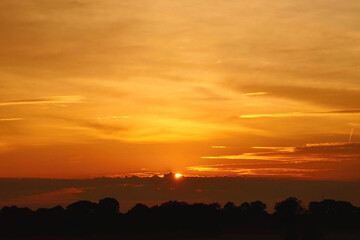 Sonnenaufgang am Horizont hinter Feld und Wald Himmel in verschiedenen Rot-, Orange- und Gelbtönen Sonne mittig halb zu sehen