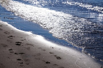 Wasserwellen am Strand bildfüllend verlaufen quer durch das Bild Sonne glitzert auf dem Wasser im Vordergrund Ufer feuchter Sand mit Fußspuren