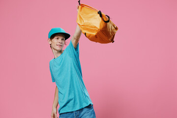 a happy, funny guy in a blue t-shirt and cap happily waves his bright backpack while standing on a pink background with empty space for advertising text