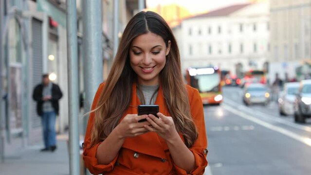 Portrait of beautiful smiling woman walking in city street texting on mobile phone with blurred background