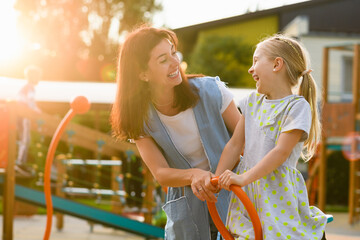Sunny family portrait of mother and daughter at playground seesaw, smiling happy mother and female...