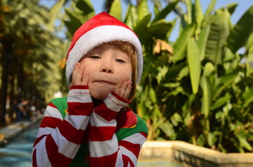 Portrait of a child in a Santa Claus hat at a tropical resort. The boy is holding his head, a lot of emotions, he is surprised, space for text. Christmas concept: waiting for a gift, family trip, 