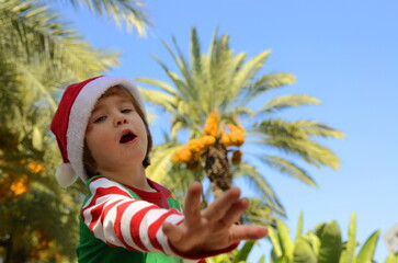 Portrait of a child in a Santa Claus hat at a tropical resort. The boy is holding his head, a lot of emotions, he is surprised, space for text. Christmas concept: waiting for a gift, family trip, 
