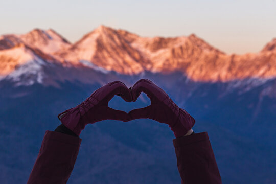 Woman making heart shape with hands over snowy mountains in the winter twilight