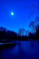 Fototapeta premium Vertical shot of a calm river surrounded by snow in a vibrant blue sky background at night
