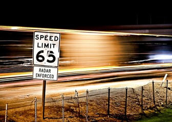 Pole with a speed limit 65 sign on the background of light trails of cars in a long exposure shot - obrazy, fototapety, plakaty
