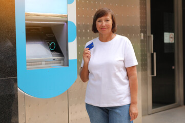 Beautiful middle aged woman is showing credit card near bank cash machine outdoors. Mature female...