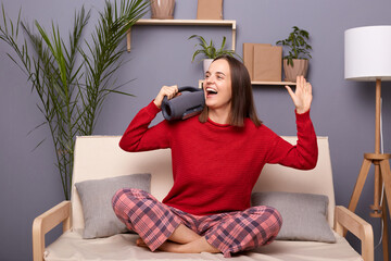 Indoor shot of happy festive brown-haired female wearing red sweater and checkered pants sitting on cough in room at home, holding music column, enjoying songs, dancing.
