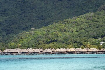 Bungalows on the water surface in Moorea Island with a green-covered hill on the background