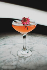 Tall orange cocktail with edible flower garnish sitting in couple glass on marble table.