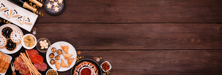 New Years Eve theme appetizer corner border. Overhead view on a dark wood banner background. Charcuterie board, champagne, clock cookies, tuxedo brie cheese and a collection of party food. Copy space.