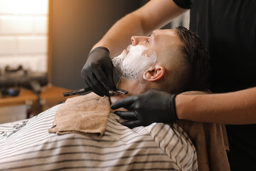 Professional barber in black gloves is shaving client with a vintage straight stainless steel razor...