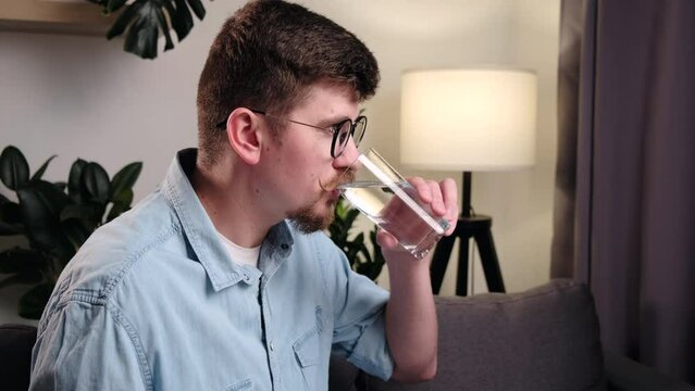 Young man in eyeglasses works at home sitting on couch, looks at computer monitor and drinking water. Handsome bearded guy drinks water from glass. Useful habits and healthy lifestyle concept