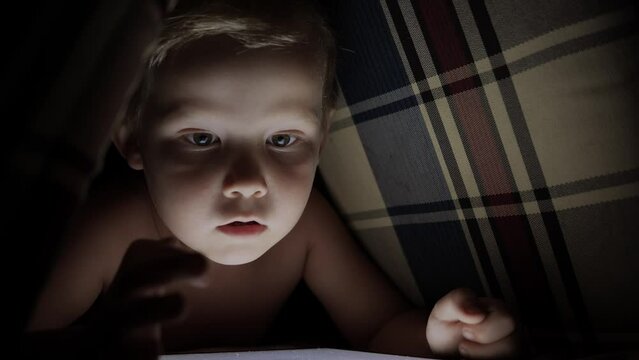 A little boy draws with his finger or watches cartoons on an electronic tablet in a pillow hut in the dark. The concept of children's interesting games and secrets. High quality 4k footage