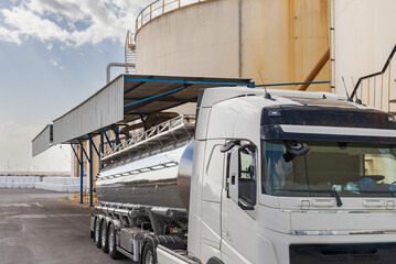 Tanker truck loading liquid next to some large tanks, with the loading area covered to protect it from the rain.