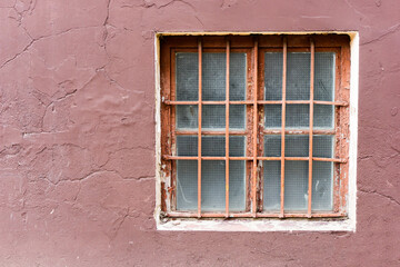 Fototapeta na wymiar Metal grate window. Behind the bars. Reinforced glass window. Peeling paint wall. Small prison window texture. Security grid pattern.