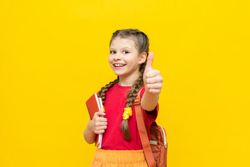 A schoolgirl is preparing a satchel for school. A little girl gives a thumbs up and collects notebooks, textbooks for the school day on a yellow isolated background. Preparation for exams.