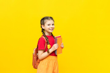 A schoolgirl with a backpack and notebooks is preparing for additional training courses. A beautiful little girl is going to school on a yellow isolated background. - obrazy, fototapety, plakaty