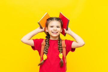 A schoolgirl is preparing a satchel for school. A little girl collects notebooks and textbooks for the school day on a yellow isolated background. Preparation for exams.