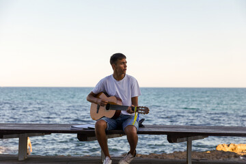 A young stylish guy plays the guitar on the embankment by the sea, the concept of music and relaxation