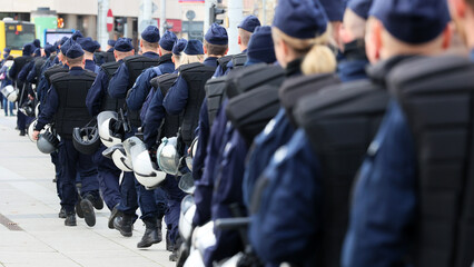 Policjanci prewencji w mundurach i tarczami idą na akcję w szeregu. 