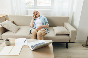 a happy, relaxed elderly woman with short white hair is sitting on a cozy sofa in an apartment and talking on a smartphone stylishly dressed in a light shirt and with glasses on her face